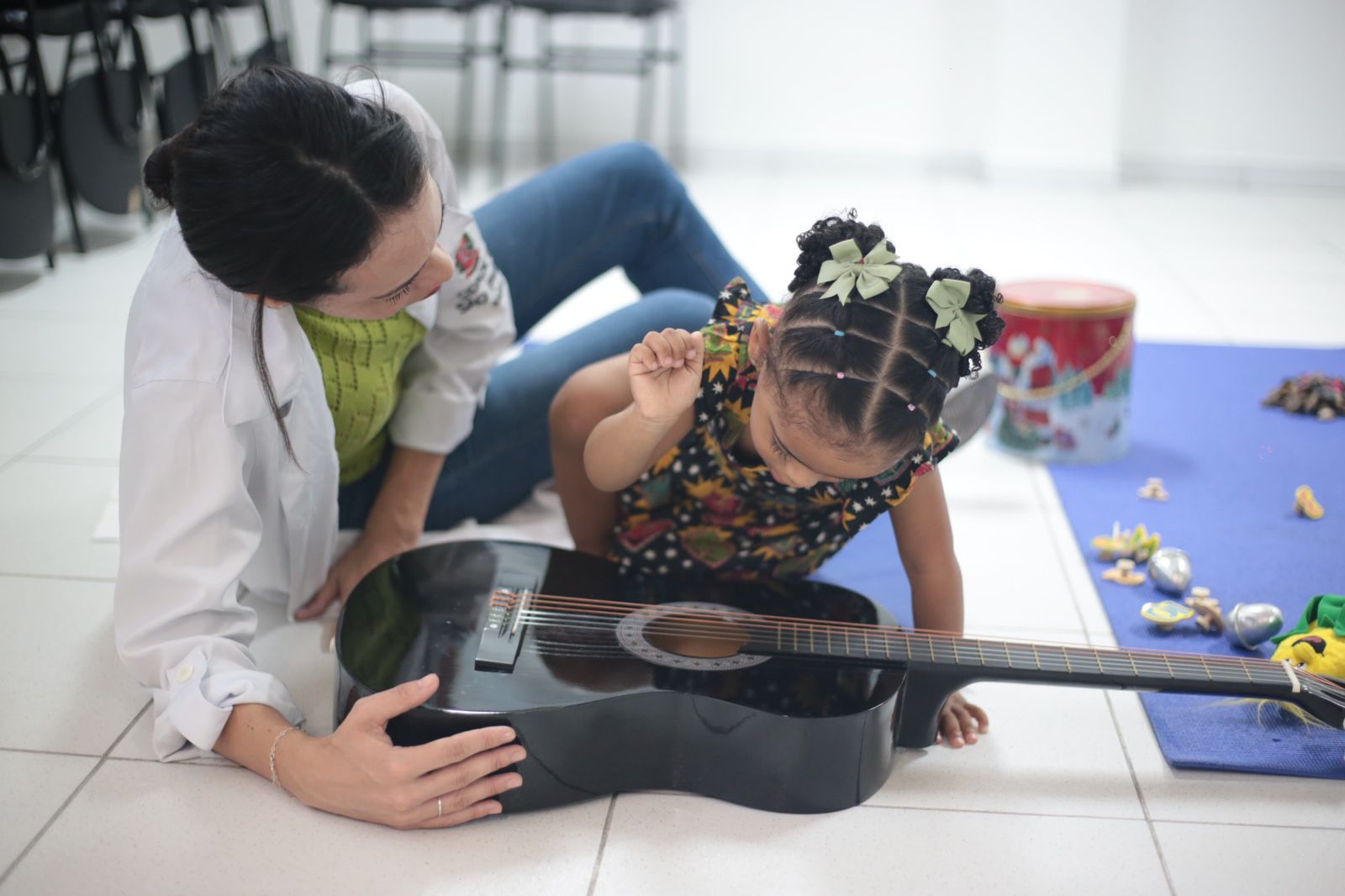 Na imagem, há uma professora e uma criança mexendo no violão, ambas sentadas no chão com brinquedos espalhados ao redor